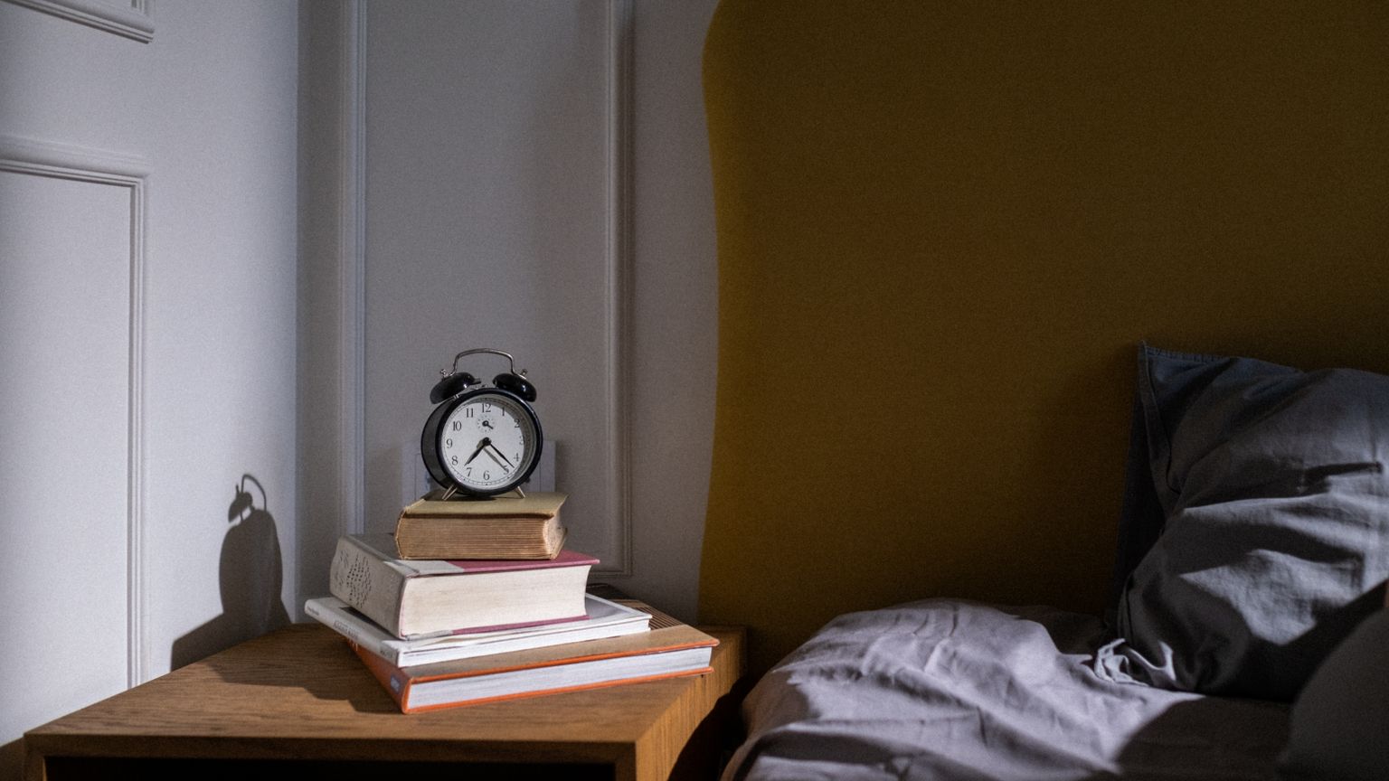 An alarm clock on a stack of books beside a bed in early light