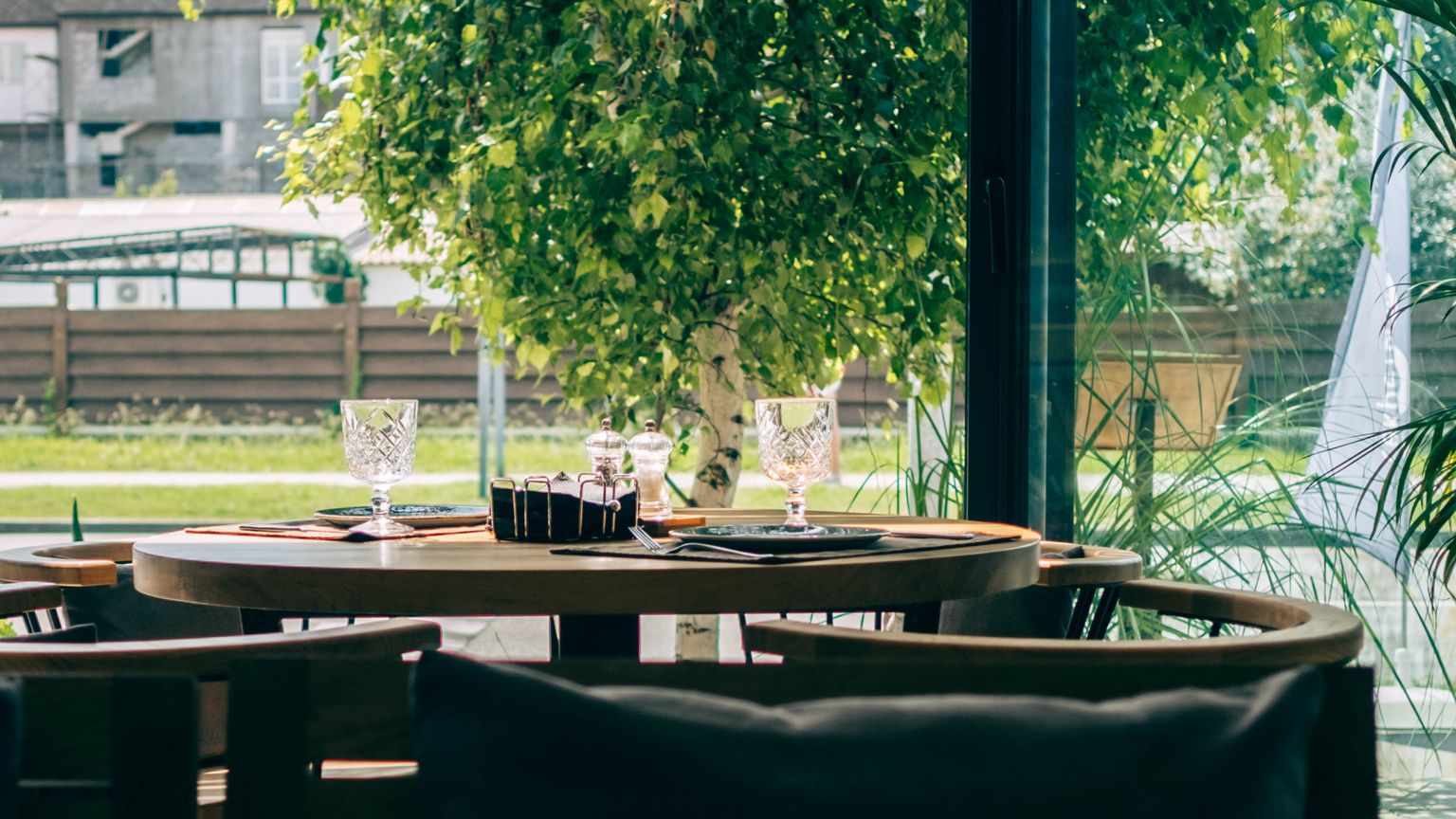 A calm restaurant table by a large window with green trees outside at midday